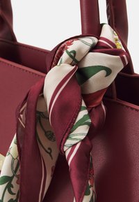 Bordó kožená kabelka s dvojitými uchy a uvázaným hedvábným šátkem s květinovým vzorem v krémových, zelených a červených odstínech.