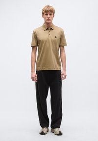 Polo tričko z ľahkej hnedej bavlny s trojmiestnou légou a malým čiernym logom, v kombinácii s tmavosivými nohavicami so širokými nohavicami a béžovými teniskami.