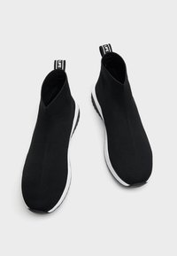 Scarpe sportive nere senza lacci con tomaia in maglia, suola bianca e linguette di trazione che presentano un logo a contrasto sul tallone.