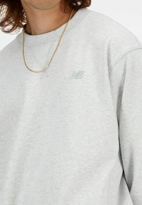 Sudadera gris de algodón, con un sutil patrón texturizado, escote en V y un logo bordado en el pecho. Collar de cadena visible.