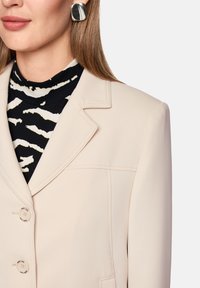 Beige blazer with notch lapels, two front buttons, and stitching details, worn over a black top with a white abstract pattern. Silver square earrings.