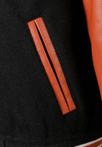 Schwarze Stoffjacke mit orangefarbenen Lederakzenten. Verfügt über eine seitliche Einschubtasche und einen gerippten Saum. Texturiertes, wollähnliches Material mit glattem Leder.