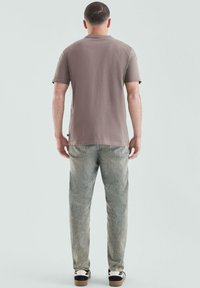 Graues Baumwoll-T-Shirt mit kurzen Ärmeln, kombiniert mit verwaschenen Jeans. Das Model trägt schwarz-weiße Sneaker und steht vor einem hellen Hintergrund.