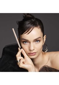 Crayon à maquillage dans un tube beige clair tenu par un mannequin. Elle a des cheveux noirs raides, une boucle d'oreille, et porte un top en tissu transparent noir.