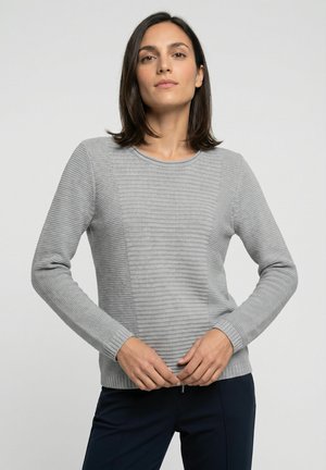 Vrouw met steil donker haar draagt een lichtgrijze, gestructureerde gebreide trui en donkere broek, staand tegen een effen lichte achtergrond.