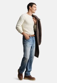 Roomkleurige kabelgebreide trui, lichtblauwe jeans, bruine leren schoenen en een donkerbruine getextureerde jas, nonchalant over één schouder gedragen.