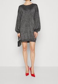 Abito in maglia nera scintillante con maniche oversize e orlo sopra al ginocchio, abbinato a scarpe rosse con punta e a una piccola borsa nera.