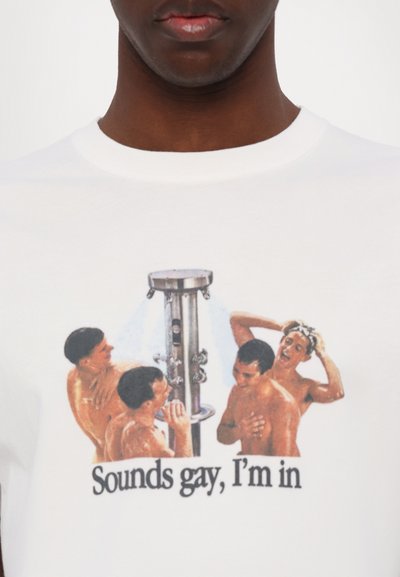 Le t-shirt montre quatre hommes torse nu prenant une douche ensemble autour d'une pomme de douche commune avec le texte "Sounds gay, I'm in" en dessous.