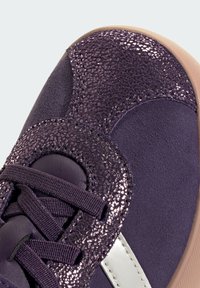 Fialová sneaker s texturovaným metalickým povrchem, hladkou semišovou částí a tónovým šněrováním; zahrnuje kontrastní světlou podrážku.