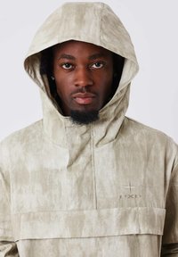 Giacca a pullover color khaki con grande cappuccio, tessuto testurizzato, pannello orizzontale frontale e logo ricamato sul petto. Finitura liscia e opaca.