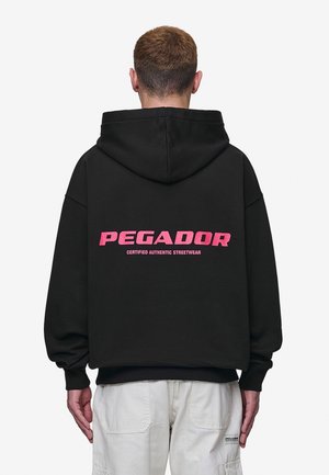 Mand iført sort hættetrøje med pink tekst "PEGADOR Certified Authentic Streetwear" på ryggen, kombineret med lyse cargobukser.