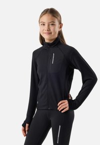 Jeune femme portant une veste noire zippée intégrale et des leggings avec des trous pour les pouces, debout avec une main sur la hanche devant un fond uni.