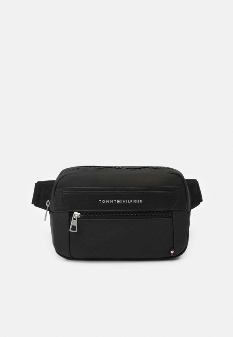Riñonera negra hecha de nylon resistente. Cuenta con un bolsillo frontal con cremallera, una correa ajustable y un logo en la parte delantera. Diseño rectangular simple.