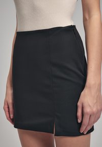 Černá mini sukně s hladkou texturou, která má boční zip a skromný přední rozparek pro přidaný detail.