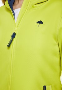 Giacca zip-up gialla brillante realizzata in materiale texturizzato, con un piccolo logo di ombrello nero sul petto e un rivestimento blu navy all'interno del cappuccio.