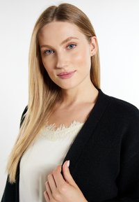 Schwarzer Cardigan mit einem strukturierten Strickdesign; getragen über einem cremefarbenen Oberteil mit einem Spitzenbesatz am Ausschnitt. Modell hält den Cardigan und hat lange Haare.