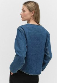 Veste en denim avec des manches longues et larges, un col rond et une coupe courte. La couleur est bleu moyen avec des détails de couture visibles.