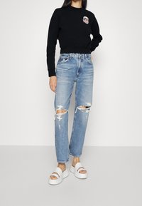 Sudadera negra recortada con un gráfico en el frente. Vaqueros de tiro alto azul claro con rodillas rasgadas. Sandalias blancas de dos correas completan el conjunto.