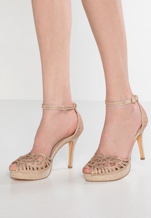 Sandały na obcasie z odkrytymi palcami, ozdobione designerskim, ciętym wzorem. Wykonane z błyszczącego złotego materiału, z cienkim paskiem na kostkę i szpilką.