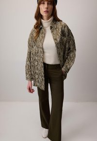 Camicia a fantasia verde oliva sopra un dolcevita crema, abbinata a pantaloni verdi scuri e sneakers bianche; materiali texturizzati e vestibilità rilassata.