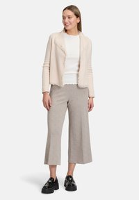 Cardigan en tricot beige clair avec des bords effilochés, porté au-dessus d'un haut blanc et d'un pantalon large gris clair. Des mocassins à plateforme noirs complètent la tenue.