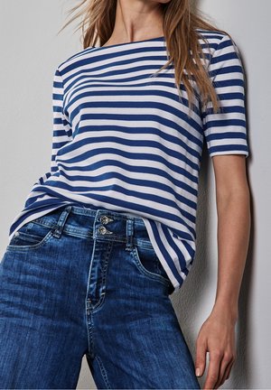 Frau trägt ein blau-weiß gestreiftes kurzärmeliges Oberteil, das in eine hochtaillierte dunkelblaue Jeans gesteckt ist, vor einer hellen Wand.