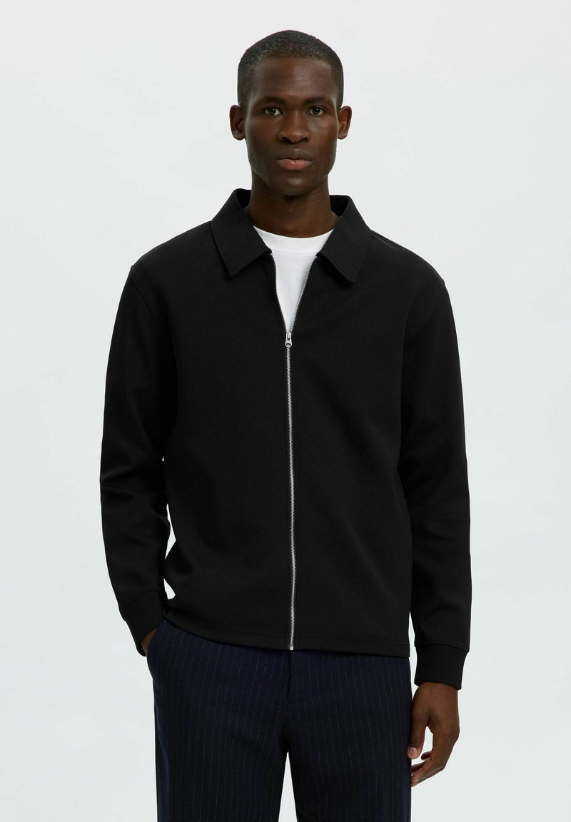 Jeune homme portant une veste noire zippée par-dessus une chemise blanche et un pantalon sombre à fines rayures, debout devant un fond clair uni.