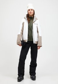 Weiße und beige isolierte Jacke mit Kapuze, grünes Basisschicht, schwarze Hose und Stiefel. Modell hält Skibrille, minimales Muster.