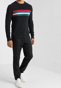 Pull bleu marine avec des rayures horizontales rouges, vertes, bleu clair et jaunes ; associé à un pantalon gris foncé texturé et des baskets blanches.