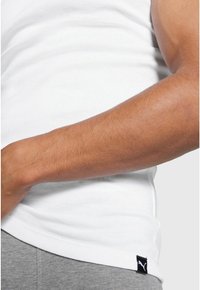White sleeveless shirt with a soft texture, featuring a black Puma logo tag on the left side, paired with grey shorts.