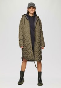Olivgrüner, gesteppter Puffer-Mantel mit Kapuze, ausgestattet mit einem Reißverschluss und seitlichen Schlitzen. Trägt man über einem schwarzen Hoodie und kombiniert mit schwarzen Stiefeletten.