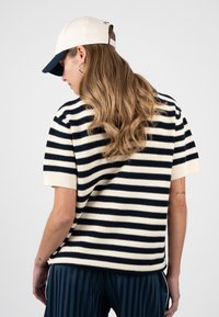 Femme aux longs cheveux ondulés portant un haut à manches courtes rayé noir et blanc, un pantalon rayé bleu marine, une casquette de baseball blanche et bleu marine, de dos.