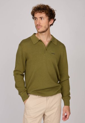 Polo en tricot vert olive avec col, manches longues et coupe décontractée, arborant un petit logo sur le côté gauche. Porté avec un pantalon beige.