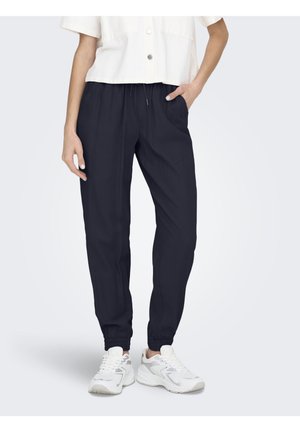 Persona con pantalones de chándal azul marino con puños elásticos y zapatillas blancas, de pie con una mano en el bolsillo y camiseta blanca corta.