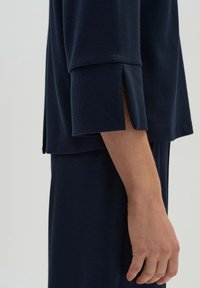 Suéter azul marino con tela texturizada, mangas hasta el codo y aberturas laterales. Mano visible sosteniendo la prenda.