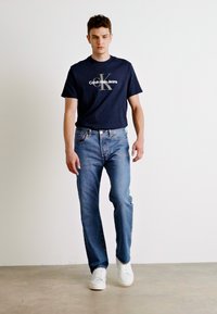 Jeans de mezclilla azules combinados con una camiseta de algodón azul marino que presenta un logo gráfico, usados con zapatillas blancas sobre un fondo neutro.