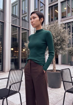 Jeune femme aux cheveux courts, en pull vert et pantalon marron, se tient dans une cour extérieure moderne avec des chaises noires et de grands arbres en pot.