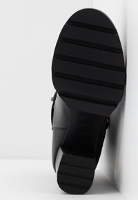 Semelle de bottes noire avec des motifs de bandes horizontales larges, inclinée contre un mur et un sol blancs.