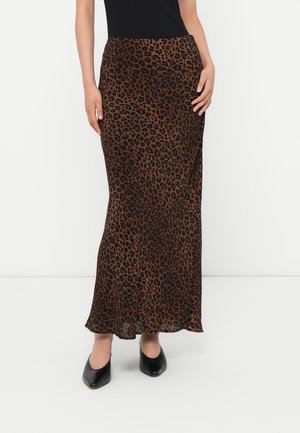 Vrouw in een lange rok met een bruin en zwart luipaardprint, zwart topje en zwarte puntige schoenen, staande tegen een witte achtergrond.