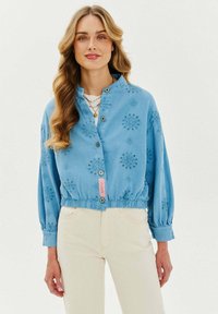 Světlemodrá košilová bunda s elastickým lemem, vyšívanými květinovými vzory a stojáčkovým límcem. Má dva knoflíky a růžovou štítkovou nášivku.