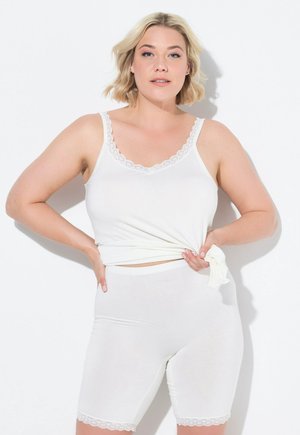 Blondine met een wit kanten topje en short, trekt het topje iets naar de zijkant, staand tegen een witte achtergrond.