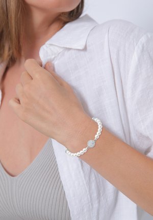 Perlenarmband mit weißen Perlen, silbernen Akzenten und einem dekorativen, runden Anhänger mit Strasssteinen. Am Handgelenk getragen.