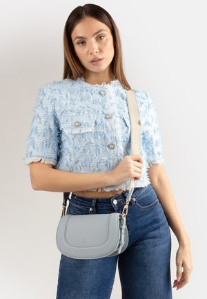 Donna che indossa una giacca corta a maniche corte di colore azzurro chiaro con trama e jeans blu scuro, tiene una borsa a tracolla grigio chiaro con una tracolla larga.