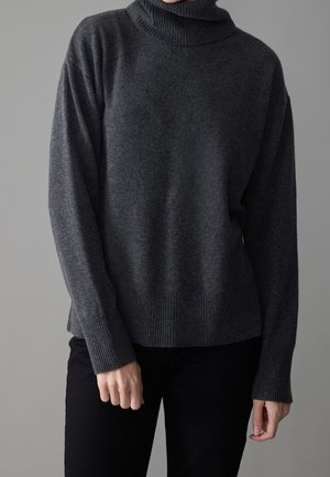Personne portant un pull à col roulé gris foncé à manches longues et un pantalon noir, debout devant un fond gris uni.