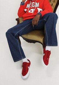 Persona sentada en un sillón vintage, vistiendo una sudadera roja, jeans azules, calcetines blancos y zapatillas rojas con detalles de estrellas blancas.