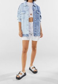 Džínová bunda v přechodu od světle modré k střední modré, oversized střih. Bílý crop top s výřezem, spárovaný s bílými šortkami a černými platformovými sandály.