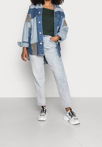 Jeans-Übergrößenjacke mit Patchwork-Design in Blau und Grau, getragen über einem grünen Oberteil und kombiniert mit hellblauen Jeans und schwarz-weißen Turnschuhen.