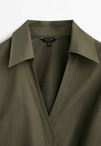 Chemise vert olive avec un col classique et des manches longues. Confectionnée en tissu lisse, elle présente un design croisé et des détails de couture subtils.