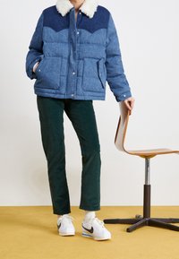 Une personne portant une doudoune bleue avec un col en fourrure blanche, un pantalon vert foncé et des sneakers Nike blanches, tenant le dossier d'une chaise en bois.