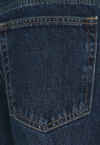 Vaqueros de mezclilla azul oscuro con un bolsillo trasero con costuras tonales y una textura suave, destacando los detalles del tejido y la construcción.